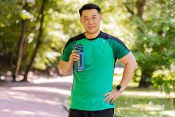Sporty athletic thirsty guy Chinese man runner jogger drinking water after workout in park outdoors...