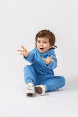Question, outrage, emotionality, expressiveness. A curly-haired toddler of 3 years old is sitting on the floor and spreading his arms in a blue tracksuit on a white background. Vertical photo. 