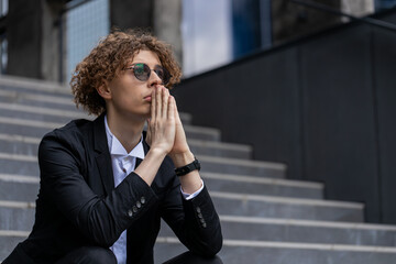 Fototapeta premium Nervous manager sitting on office steps. Pensive businessman in suit with hands joined making company decision. Successful CEO expert thinking silent break. Corporate leader pondering in downtown.