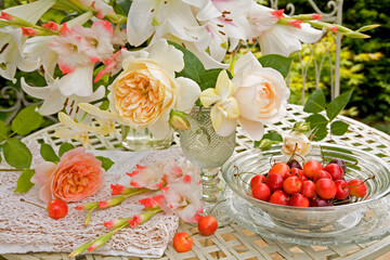 White roses, lilies and gladioli in glass vases with a bowl of cherries on  a table outdoors
