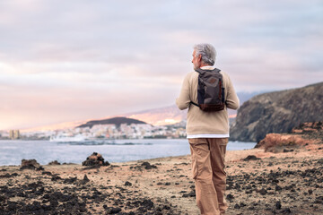 Back view of senior man walking on the cliff wearing backpack enjoying sea excursion at sunset light. Active white-haired retired man in sea vacation, healthy lifestyle concept