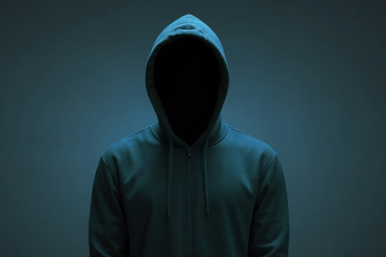 Hacker man is wearing a hoodie and is standing in front of a dark blue background. Concept of mystery and anonymity, as the man's face is hidden behind the hood