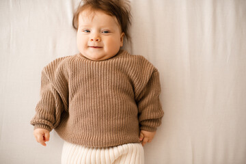 Fototapeta premium Smiling baby girl 5-6 months old lying in bed on white blanket wearing knitted sweater close up. Childhood.
