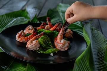 Children's hands take a large tiger shrimp lying on a black plate among tropical green banana leaves