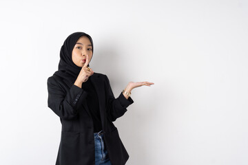 Asian moslem female with black hijab and suit put finger in front of her mouth telling quite sign with hands pointing copy space and face looking to camera