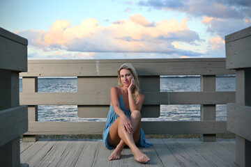 Sexy blonde woman sitting in blue dress on dock at sunset
