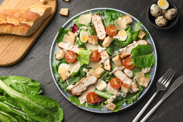 Tasty Caesar salad with chicken and cutlery on dark gray textured table, flat lay