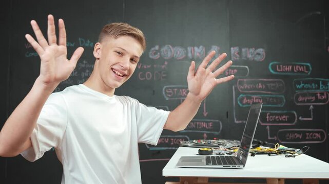 Side view of young boy programming code while looking at camera and greeting. high school student coding system while sitting at STEM classroom with electronic equipment scatter on table. Edification