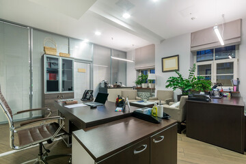 A brightly lit office with dark furniture. A visitor area with soft light sofas. Green plants on the windows..