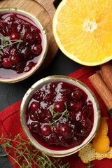 Tasty cranberry sauce in bowls, orange, thyme and cinnamon on black table, top view