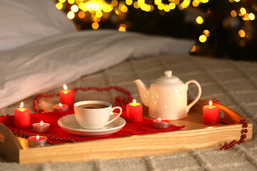 Aromatic tea in cup, teapot and burning candles on bed against blurred Christmas lights, bokeh effect