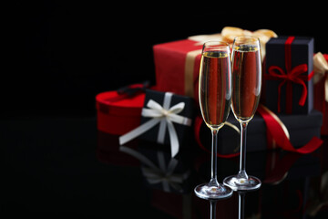 Delicious champagne in glasses and Christmas presents on black mirror surface, closeup. Space for text