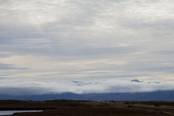 Fototapeta premium A winding road through a field, with towering, cloudy mountains looming in the distance, showcasing Iceland's raw beauty
