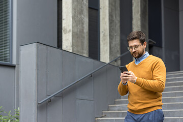 A young man businessman walks near the stairs of the office center and uses a mobile phone, dials a...