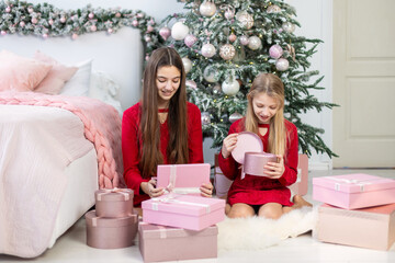 Girls opening Christmas gifts by tree. Two girls in red dresses excitedly opening pink gift boxes near a beautifully decorated Christmas tree in a cozy bedroom.