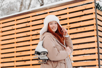 young pretty caucasian redhead smiling girl in white and beige winter clothes skating in ice rink in winter cloudy snowy day, with skates on shoulder, looking straight to camera