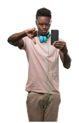 Fototapeta premium Young african american man wearing headphones and holding smartphone with angry face, negative sign showing dislike with thumbs down, rejection concept