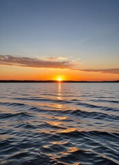 Obraz premium A scenic sunset over a calm lake, with the sun dipping below the horizon and casting warm, golden light across the rippling water