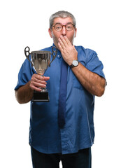 Handsome senior successful man holding trophy over isolated background cover mouth with hand...