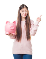 Young Chinese woman over isolated background holding piggy bank very happy pointing with hand and...