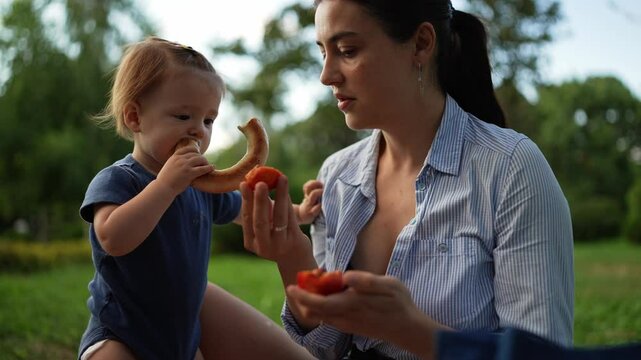 A brunette woman in a shirt offers her little daughter a piece of fruit but the child prefers a bagel during her picnic in the park in summer