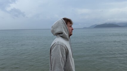 Young man shivering and trembling from cold rainy weather at summer vacation on beach, sea