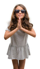 Brunette hispanic girl wearing sunglasses begging and praying with hands together with hope expression on face very emotional and worried. Asking for forgiveness. Religion concept.