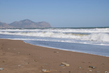 A sandy beach, large sea waves with white foam, rocks in the distance on a clear sunny day.
