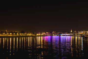 Night view of the Sava Bridge illuminated in various colors and its reflection in the river while the banks of old and new Belgrade are visible behind it