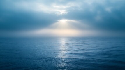 Obraz premium Sunbeams pierce through misty clouds over calm ocean.