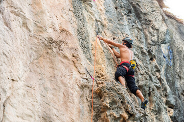 Extreme sport climbing on summer beach holiday vacation. Healthy strong Asian man enjoy outdoor active lifestyle climbing on rocky mountain with safety equipment and hanging rope at tropical island.
