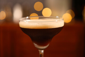 Delicious espresso martini in glass against blurred lights, closeup