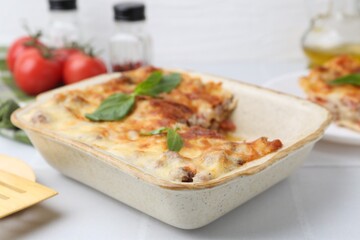 Delicious cooked lasagna in baking dish and basil on white table, closeup