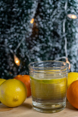 A glass of water with lemon in the foreground, surrounded by lemons and mandarins, with a Christmas tree in the background.