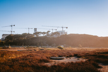 Construction cranes working on a building site on a hill at sunset, with dry grass and an abandoned boat in the foreground, in vila nova de gaia, portugal. From Estuário do Douro Local Nature Reserve.