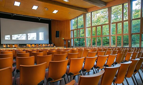 A spacious conference room with orange chairs and large windows overlooking greenery.