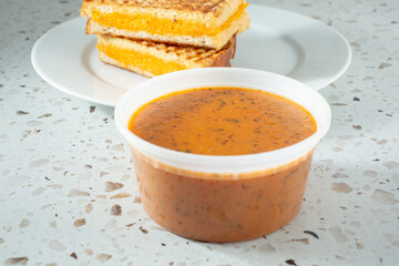 A view of a container of tomato soup, with a grilled cheese sandwich in the background.