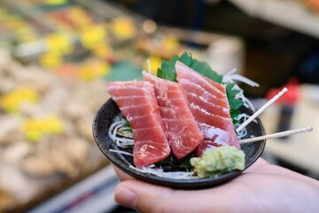 Fatty tuna sashimi slices glazed with soy sauce, and garnished with a perilla leaf and fresh wasabi, held in a woman’s hand at Tsukiji Outer Fish Market in Chuo, Tokyo, Japan 