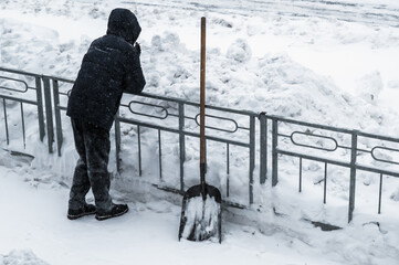 The janitor took a break from snow removal. The shovel is standing by the fence. Winter work after a snowfall. Rear view.