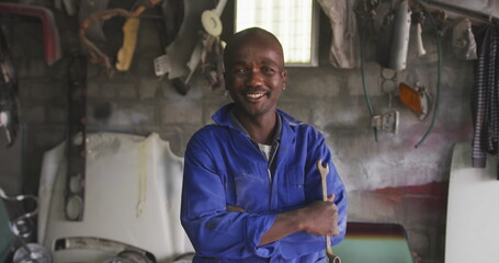 Portrait of a happy African male panel beater in a township workshop, holding a wrench, looking at c