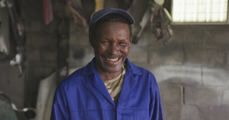 Portrait of a happy senior African male panel beater in a township workshop, wearing a cap, looking