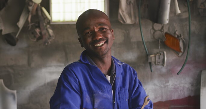 Portrait of a happy African male panel beater in a township workshop, holding a wrench, looking at c