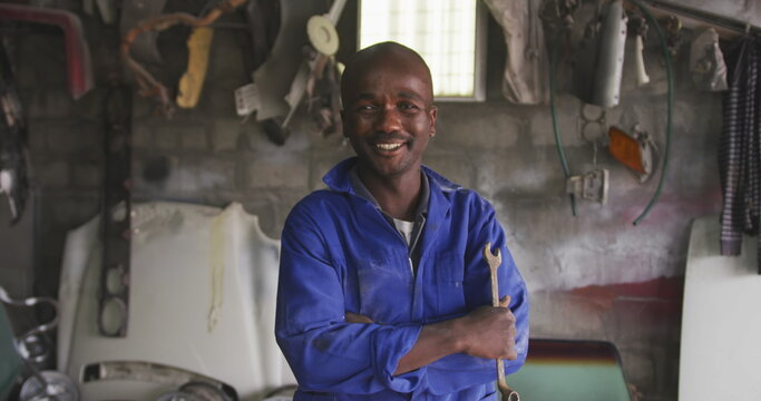 Portrait of a happy African male panel beater in a township workshop, holding a wrench, looking at c