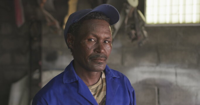 Portrait of a senior African male panel beater in a township workshop, wearing a cap, looking at cam