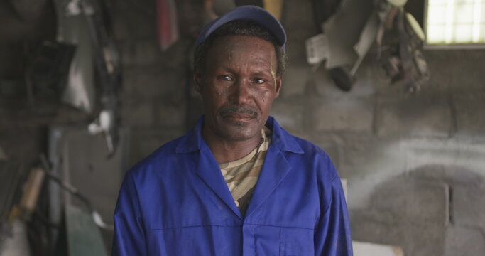 Portrait of a happy senior African male panel beater in a township workshop, wearing a cap, looking - Powered by Adobe
