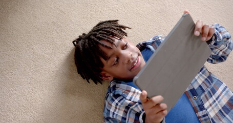 Happy african american boy using tablet lying on floor at home, slow motion - Powered by Adobe