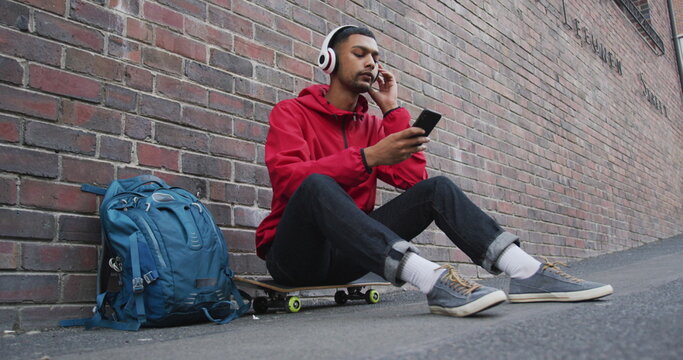 Biracial man sitting on skateboard and listening to music in the street - Powered by Adobe