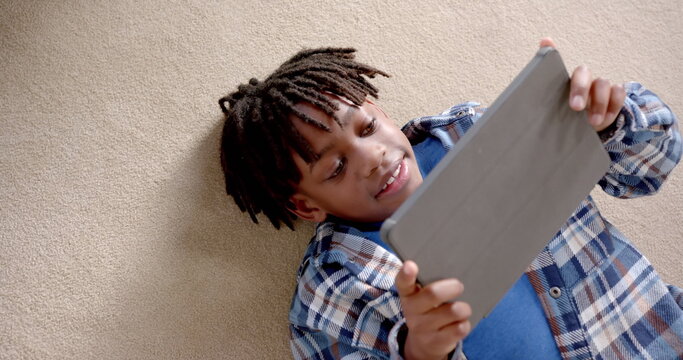 Happy african american boy using tablet lying on floor at home, slow motion - Powered by Adobe