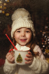 A little girl in a white hat and a sweater looks through the window, snow-covered glass. In the hands of a mug with a Christmas tree. Christmas. New Year.