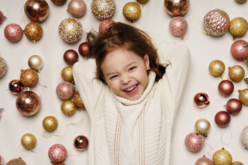 A little girl is lying on her back and laughing. Top view, portrait. Christmas balls on the background. Christmas. New Year.